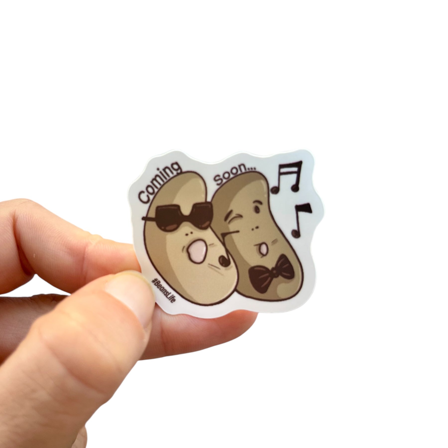 Hand holding a cartoon-style 'Coming Soon' sticker with musical notes on a white background